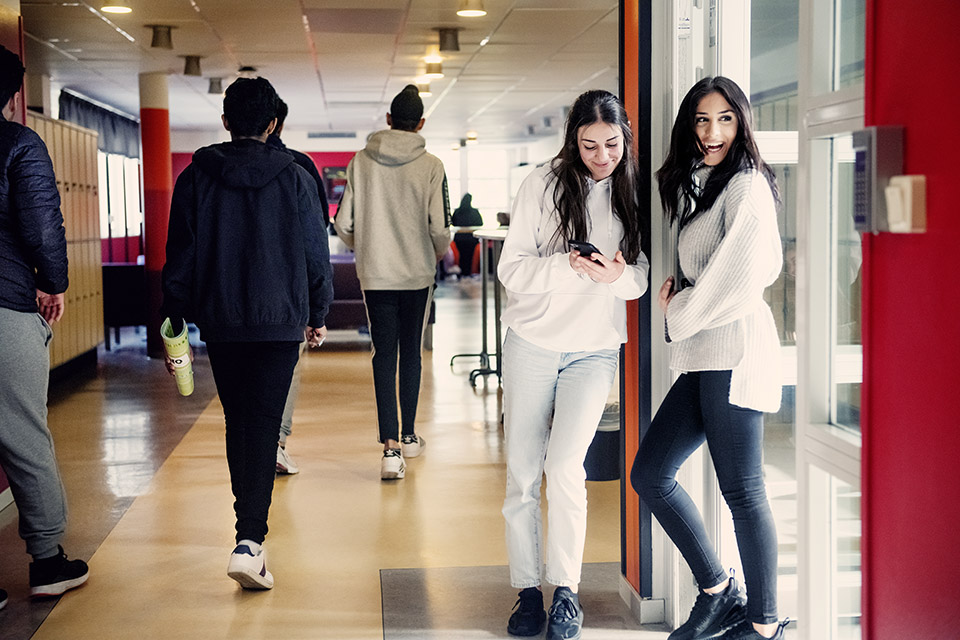 Två tjejer står vid dörren av en skolkorridor. En skrattar medan den andra kollar sin mobil.
